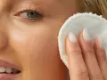 Viso di donna bionda con pelle luminosa, mentre rimuove il trucco con un dischetto. La sua pelle sembra liscia come grani di miglio bucarli.