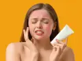 Donna con viso arrossato per eritema solare, che tiene in mano una crema. Potrebbero comparire brufoli a causa dell'irritazione.