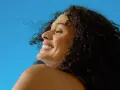 Donna sorridente con capelli ricci scuri sotto il sole, un'immagine che evoca il significato di elioterapia e benessere naturale.