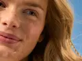 Primo piano di una donna con lentiggini e capelli biondi, sorride sotto un cielo azzurro. La sua pelle luminosa suggerisce che sa quanto dura la crema solare.