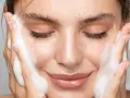 Una donna sorride mentre si lava il viso con schiuma detergente. La sua pelle luminosa e le lentiggini risaltano mentre si prende cura di sé.