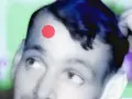 Primo piano di un volto con un punto rosso sulla fronte, come una crosta di brufolo schiacciato. Sfondo astratto verde e viola.