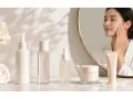 Passaggi skincare serale: flaconi, siero, crema e una donna che si accarezza il viso riflessa nello specchio.