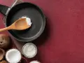Cucina con olio di cocco: una padella con un cucchiaio di olio di cocco, noci di cocco e un barattolo di olio di cocco, per scoprire i suoi molteplici usi.