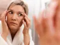 Donna in accappatoio bianco si guarda allo specchio, preoccupata per cosa fa invecchiare la pelle, toccandosi le rughe intorno agli occhi.