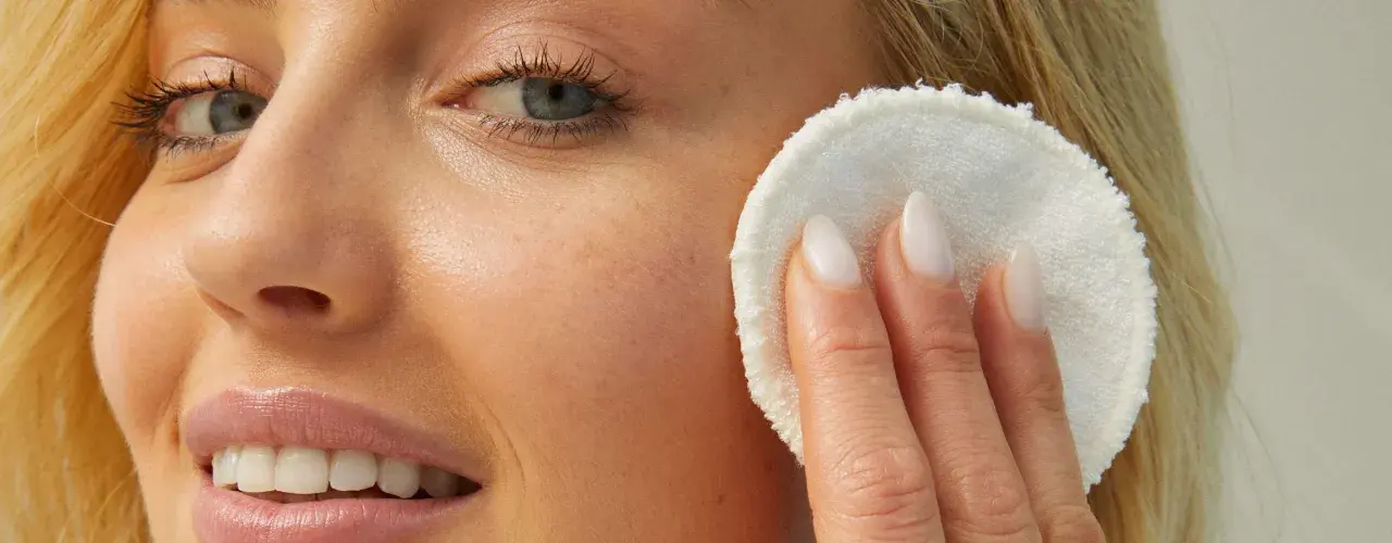 Viso di donna bionda con pelle luminosa, mentre rimuove il trucco con un dischetto. La sua pelle sembra liscia come grani di miglio bucarli.