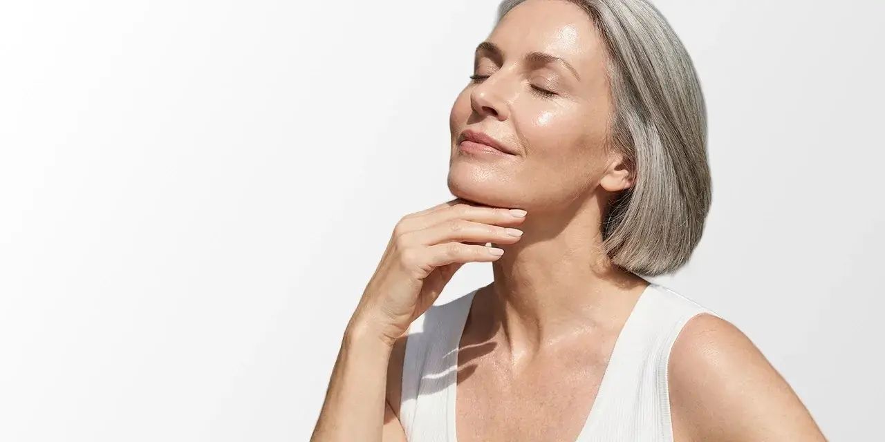 Donna con capelli grigi e pelle luminosa, che accarezza il collo. Scopri i migliori rimedi per le rughe del collo.
