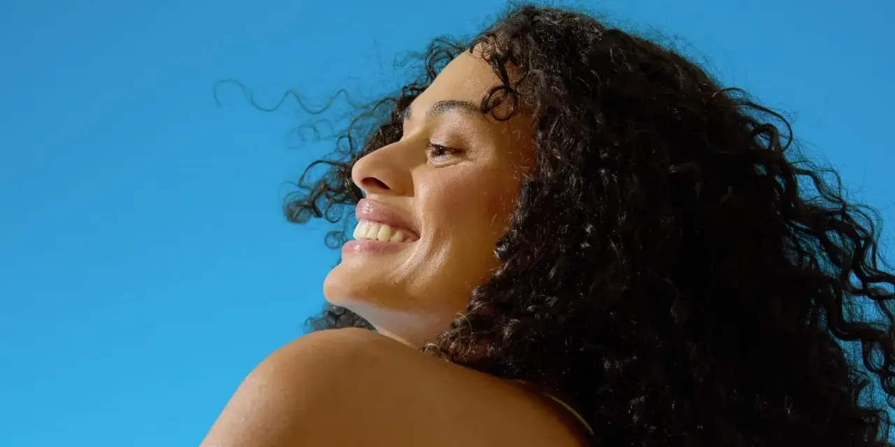 Donna sorridente con capelli ricci scuri sotto il sole, un'immagine che evoca il significato di elioterapia e benessere naturale.