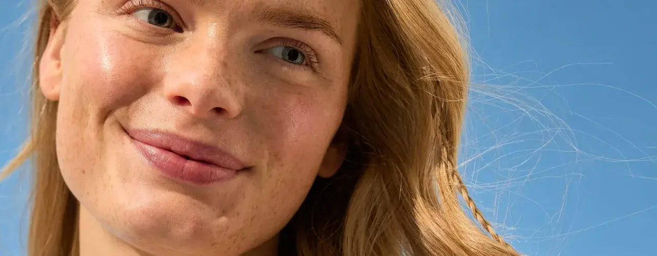 Primo piano di una donna con lentiggini e capelli biondi, sorride sotto un cielo azzurro. La sua pelle luminosa suggerisce che sa quanto dura la crema solare.