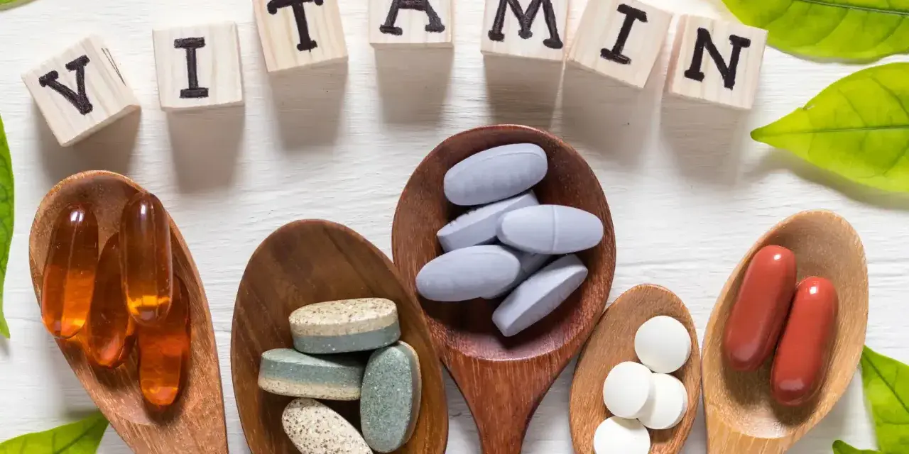 Cucchiaini di legno pieni di pillole e capsule, con le lettere che compongono la parola "VITAMIN" e foglie verdi. Tra le pillole, si nota la vitamina b6.