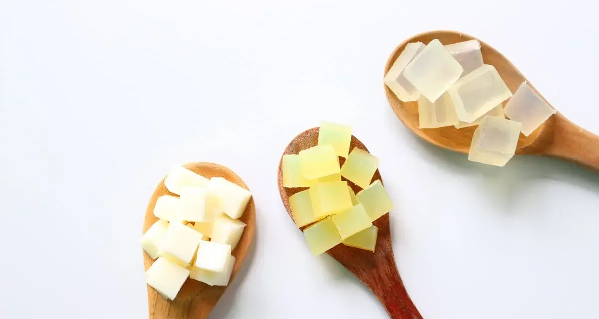 Tre cucchiai di legno con cubetti di sapone trasparente, giallo e bianco. La glicerina da dove si ricava è un ingrediente chiave per questi saponi.