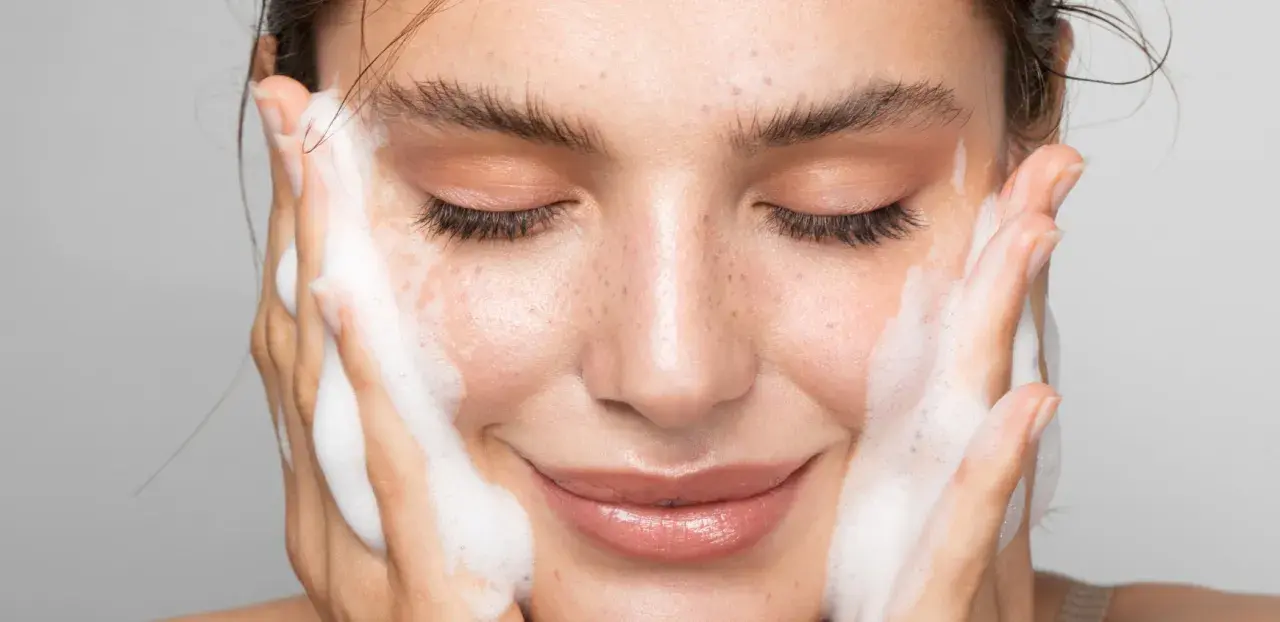 Una donna sorride mentre si lava il viso con schiuma detergente. La sua pelle luminosa e le lentiggini risaltano mentre si prende cura di sé.