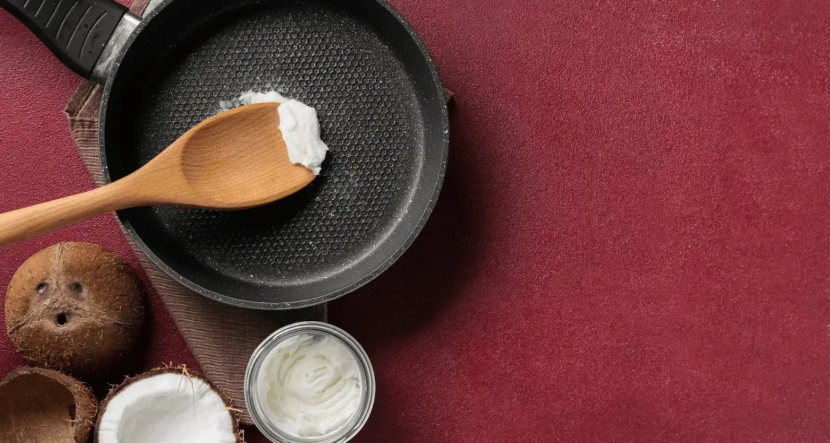 Cucina con olio di cocco: una padella con un cucchiaio di olio di cocco, noci di cocco e un barattolo di olio di cocco, per scoprire i suoi molteplici usi.