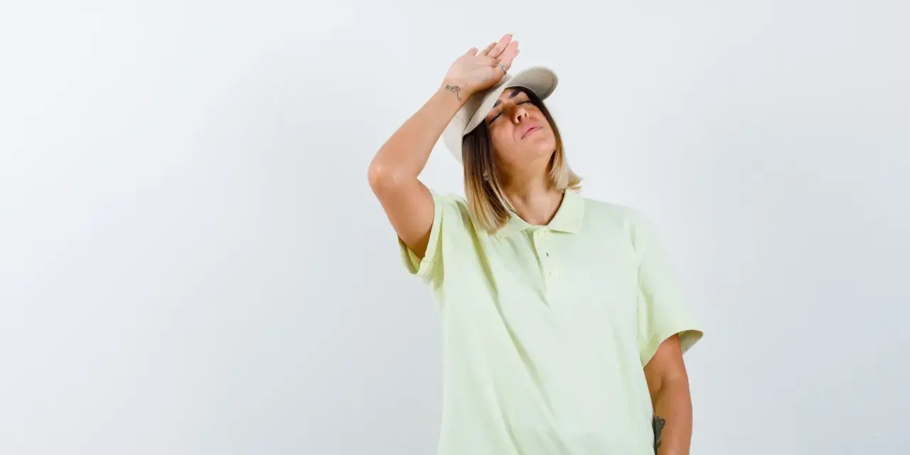 Donna con cappellino e maglietta chiara, mano sulla fronte, con espressione di chi soffre per l'insolazione quanto dura.