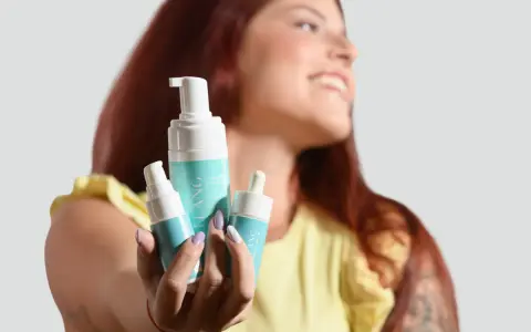 Donna sorridente mostra prodotti per skincare mattina: mousse detergente e siero.