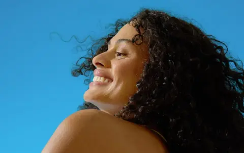 Donna sorridente con capelli ricci scuri sotto il sole, un'immagine che evoca il significato di elioterapia e benessere naturale.