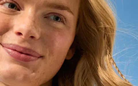 Primo piano di una donna con lentiggini e capelli biondi, sorride sotto un cielo azzurro. La sua pelle luminosa suggerisce che sa quanto dura la crema solare.