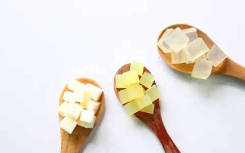 Tre cucchiai di legno con cubetti di sapone trasparente, giallo e bianco. La glicerina da dove si ricava è un ingrediente chiave per questi saponi.