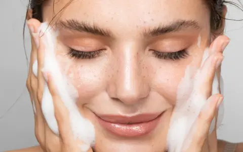 Una donna sorride mentre si lava il viso con schiuma detergente. La sua pelle luminosa e le lentiggini risaltano mentre si prende cura di sé.