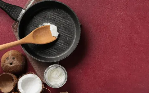 Cucina con olio di cocco: una padella con un cucchiaio di olio di cocco, noci di cocco e un barattolo di olio di cocco, per scoprire i suoi molteplici usi.