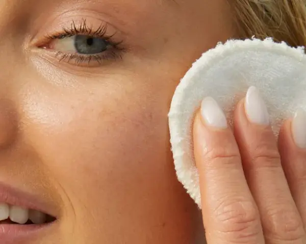 Viso di donna bionda con pelle luminosa, mentre rimuove il trucco con un dischetto. La sua pelle sembra liscia come grani di miglio bucarli.