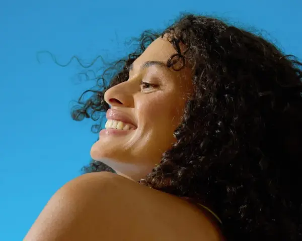 Donna sorridente con capelli ricci scuri sotto il sole, un'immagine che evoca il significato di elioterapia e benessere naturale.