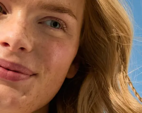 Primo piano di una donna con lentiggini e capelli biondi, sorride sotto un cielo azzurro. La sua pelle luminosa suggerisce che sa quanto dura la crema solare.