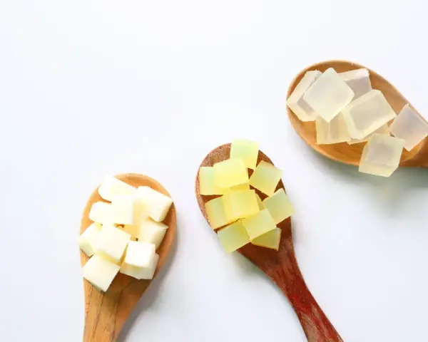Tre cucchiai di legno con cubetti di sapone trasparente, giallo e bianco. La glicerina da dove si ricava è un ingrediente chiave per questi saponi.