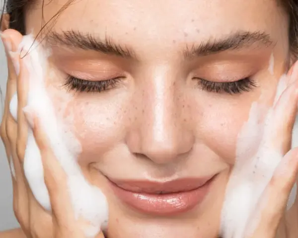 Una donna sorride mentre si lava il viso con schiuma detergente. La sua pelle luminosa e le lentiggini risaltano mentre si prende cura di sé.
