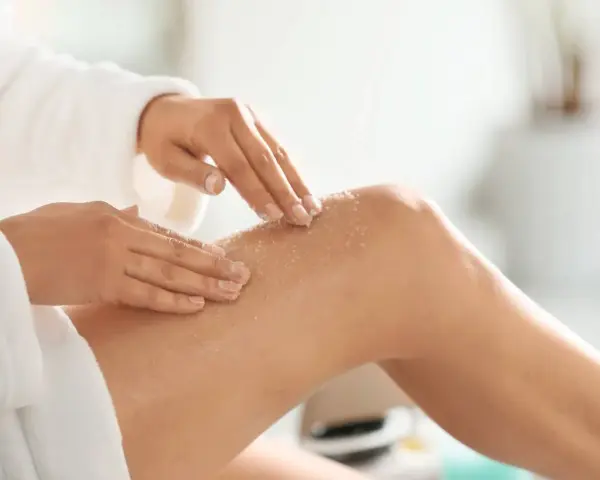 Mani che applicano body scrub sulla gamba, un gesto delicato per una pelle liscia. Scopri come si usa il body scrub per un trattamento efficace.