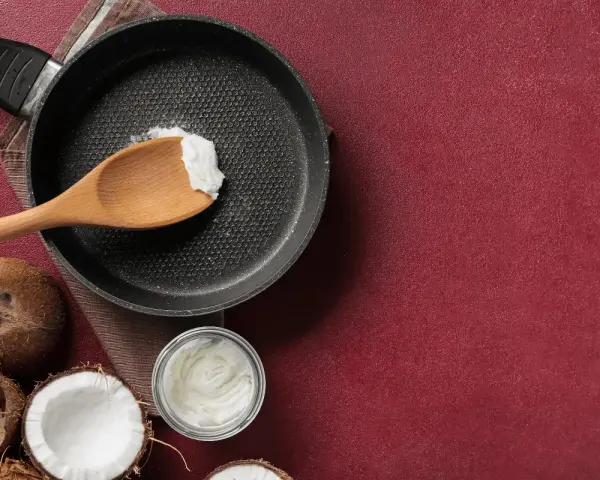 Cucina con olio di cocco: una padella con un cucchiaio di olio di cocco, noci di cocco e un barattolo di olio di cocco, per scoprire i suoi molteplici usi.