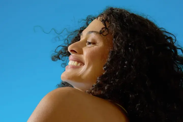 Donna sorridente con capelli ricci scuri sotto il sole, un'immagine che evoca il significato di elioterapia e benessere naturale.