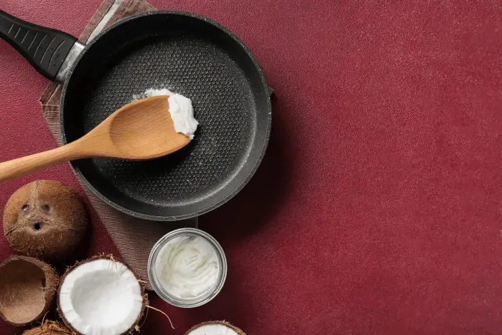 Cucina con olio di cocco: una padella con un cucchiaio di olio di cocco, noci di cocco e un barattolo di olio di cocco, per scoprire i suoi molteplici usi.