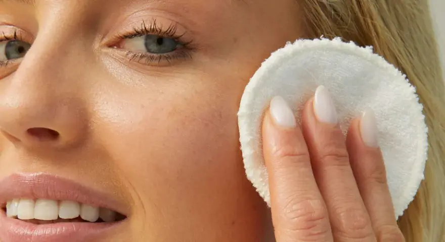 Viso di donna bionda con pelle luminosa, mentre rimuove il trucco con un dischetto. La sua pelle sembra liscia come grani di miglio bucarli.
