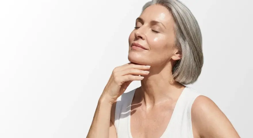 Donna con capelli grigi e pelle luminosa, che accarezza il collo. Scopri i migliori rimedi per le rughe del collo.