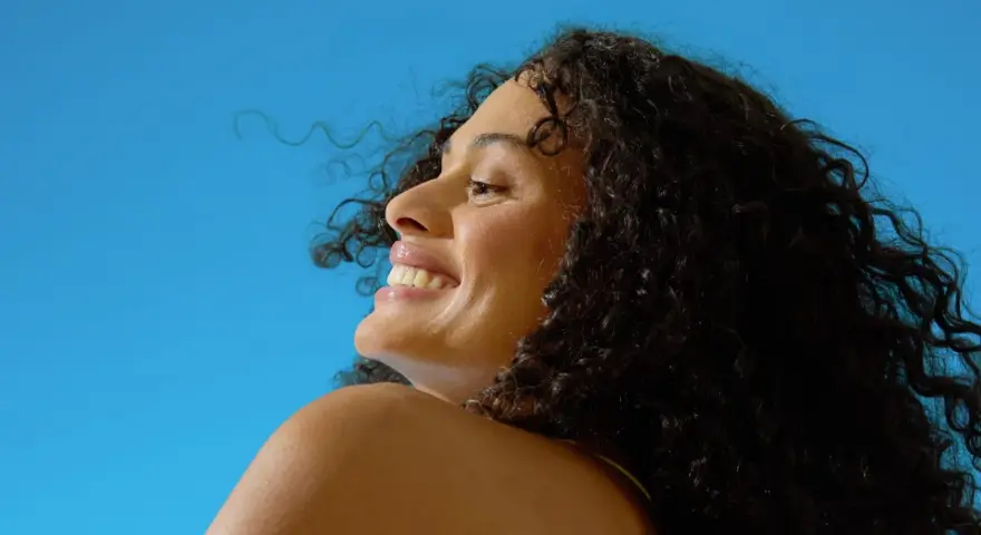 Donna sorridente con capelli ricci scuri sotto il sole, un'immagine che evoca il significato di elioterapia e benessere naturale.