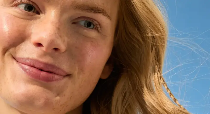 Primo piano di una donna con lentiggini e capelli biondi, sorride sotto un cielo azzurro. La sua pelle luminosa suggerisce che sa quanto dura la crema solare.