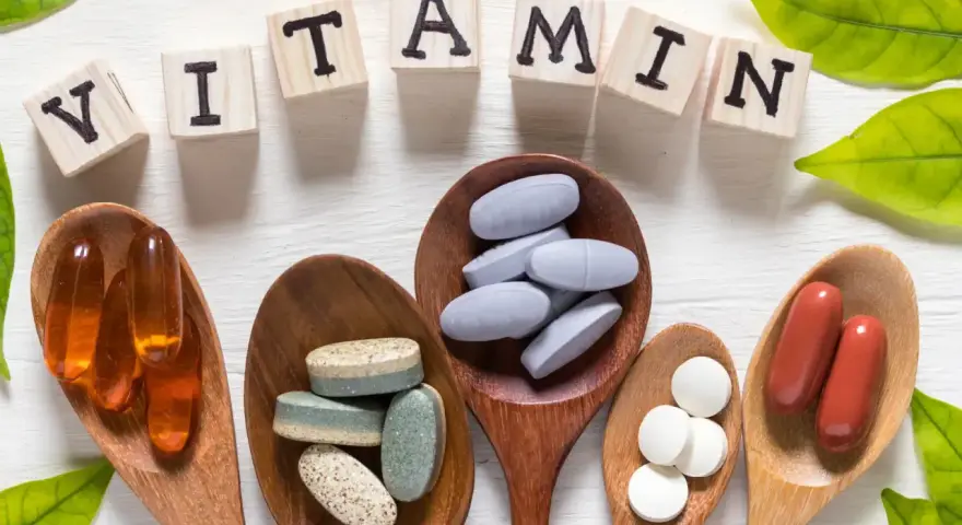 Cucchiaini di legno pieni di pillole e capsule, con le lettere che compongono la parola "VITAMIN" e foglie verdi. Tra le pillole, si nota la vitamina b6.