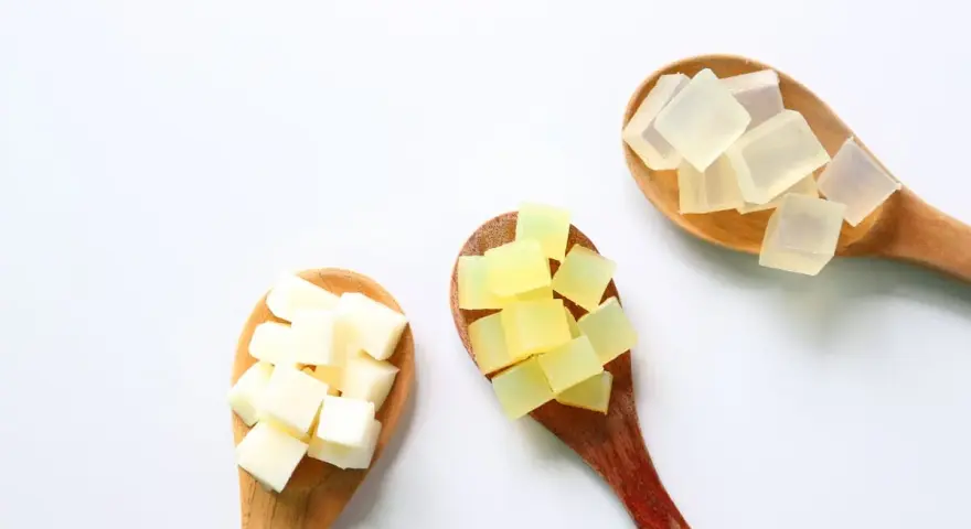 Tre cucchiai di legno con cubetti di sapone trasparente, giallo e bianco. La glicerina da dove si ricava è un ingrediente chiave per questi saponi.