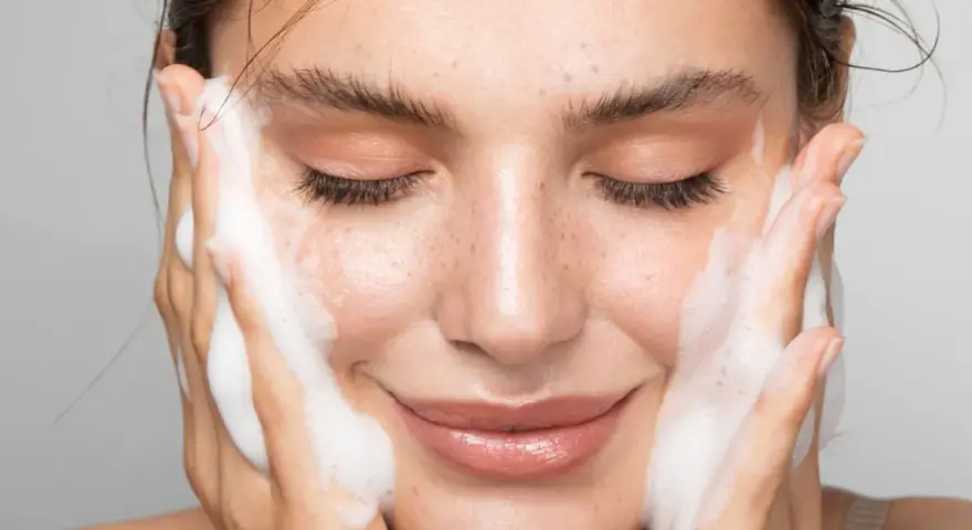 Una donna sorride mentre si lava il viso con schiuma detergente. La sua pelle luminosa e le lentiggini risaltano mentre si prende cura di sé.