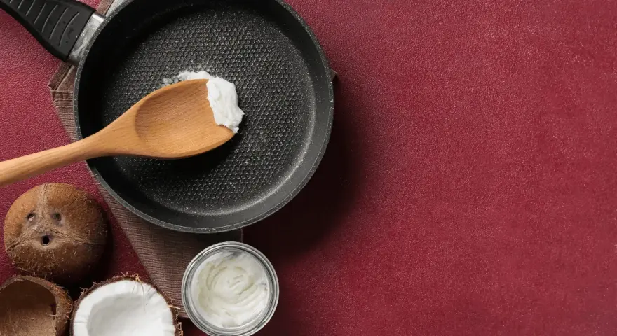 Cucina con olio di cocco: una padella con un cucchiaio di olio di cocco, noci di cocco e un barattolo di olio di cocco, per scoprire i suoi molteplici usi.