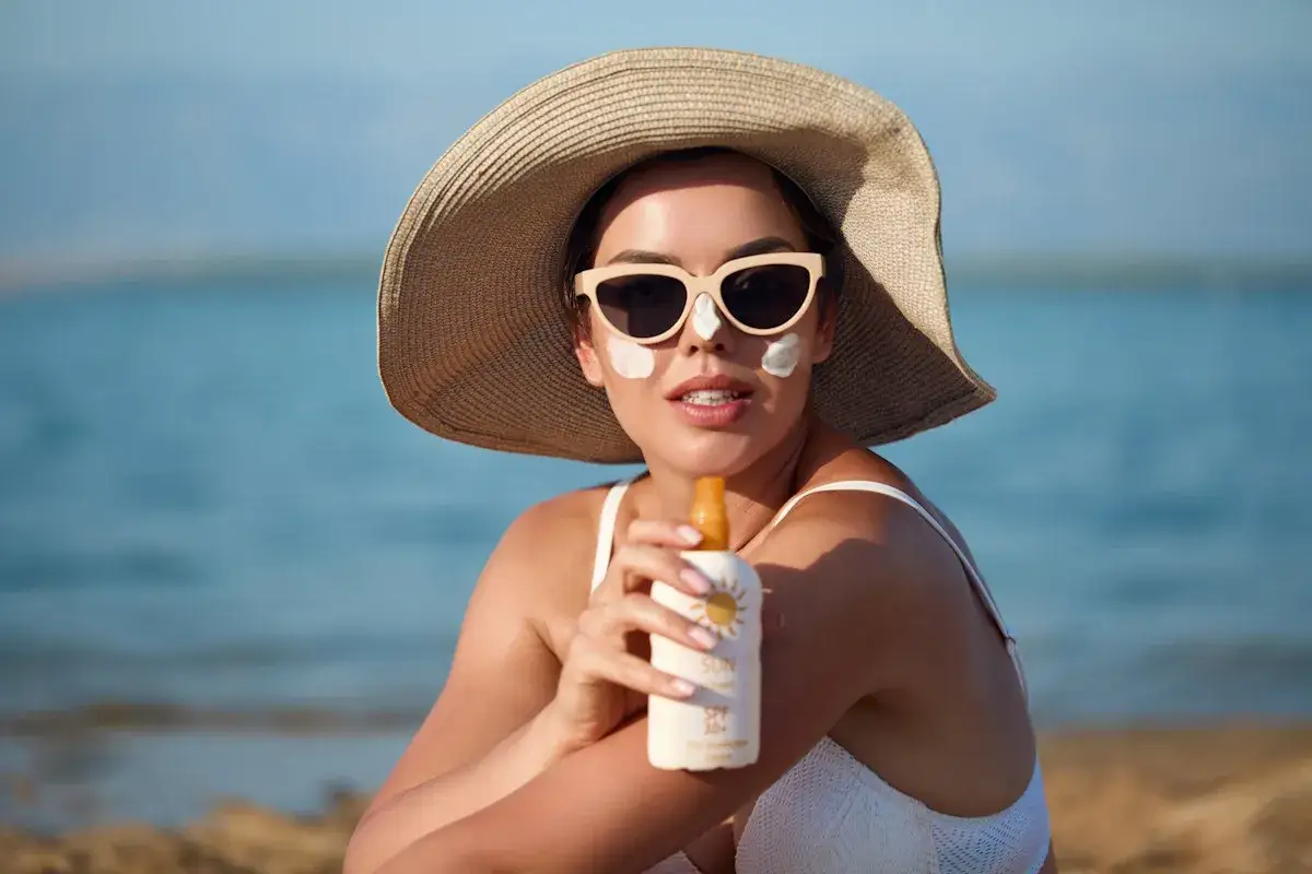 Donna con cappello e occhiali da sole, crema solare sul viso, tiene una bottiglia di protezione solare. Ricorda che prendere il sole fa bene, ma con cautela!