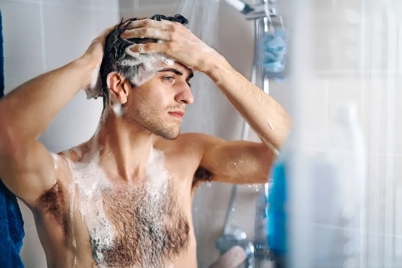 Uomo con le mani tra i capelli insaponati sotto la doccia. &Egrave; corretto fare la doccia tutti i giorni per una buona igiene.