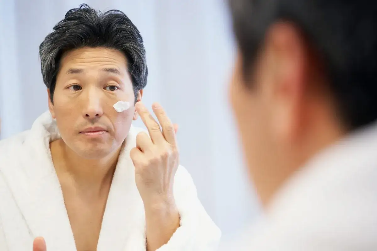 Uomo con accappatoio bianco applica crema sul viso, cercando rimedi per prurito insopportabile.