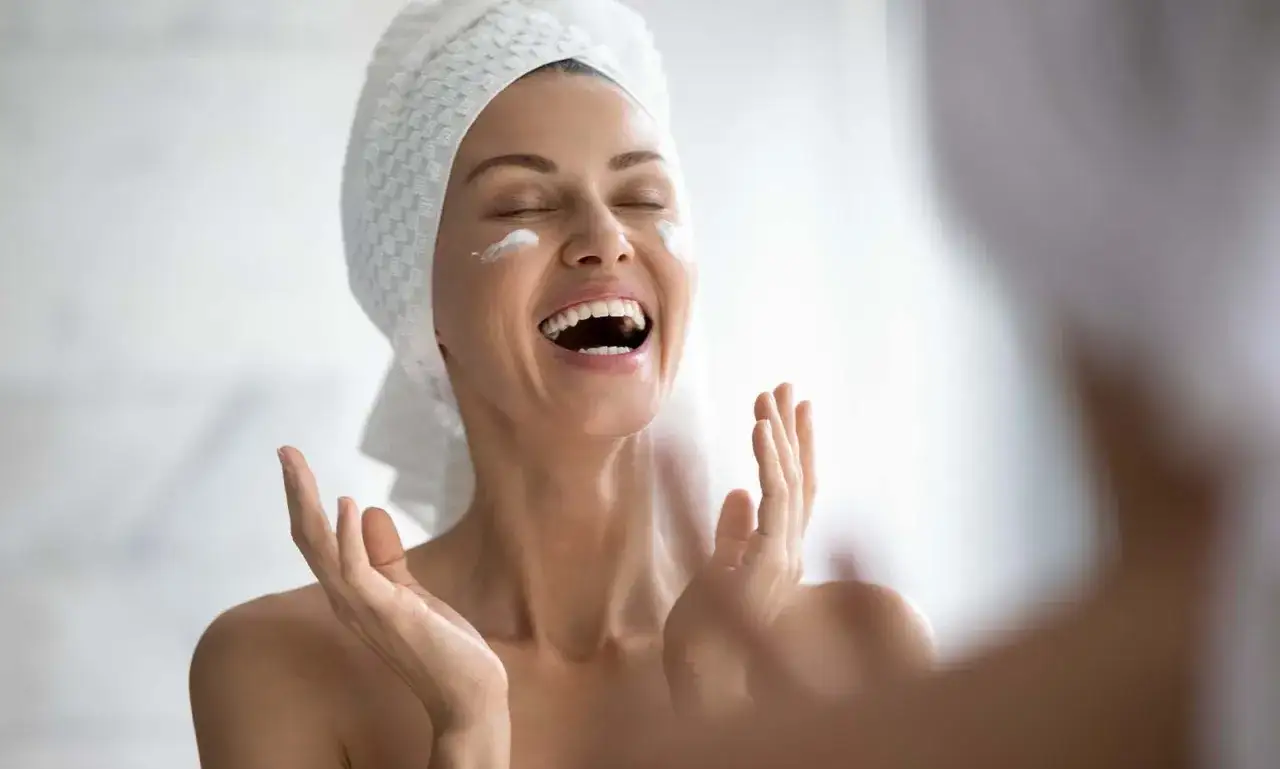 Donna con asciugamano in testa applica crema sul viso, ridendo felice. Passaggi skincare serale.