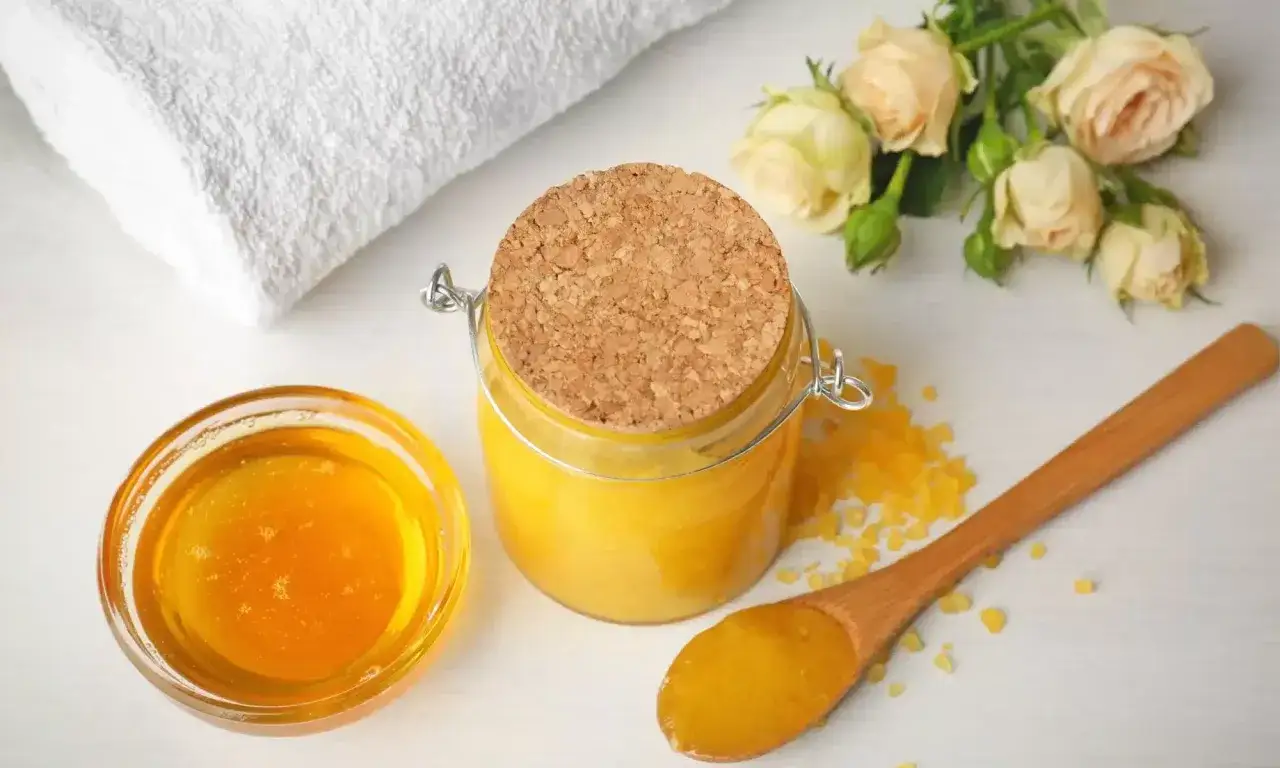 Scrub miele e zucchero fatto in casa, con un cucchiaio di legno, un vasetto di vetro, un fiorellino e un asciugamano bianco.