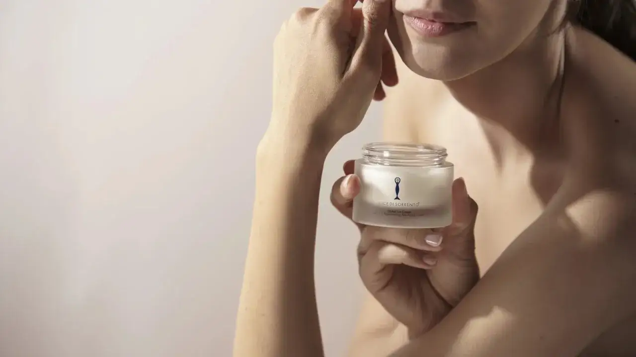 Mani delicate che accarezzano il viso, tenendo un vasetto di crema. La bellezza a 20 anni è un tesoro da custodire con cura, come le collane di Venere.