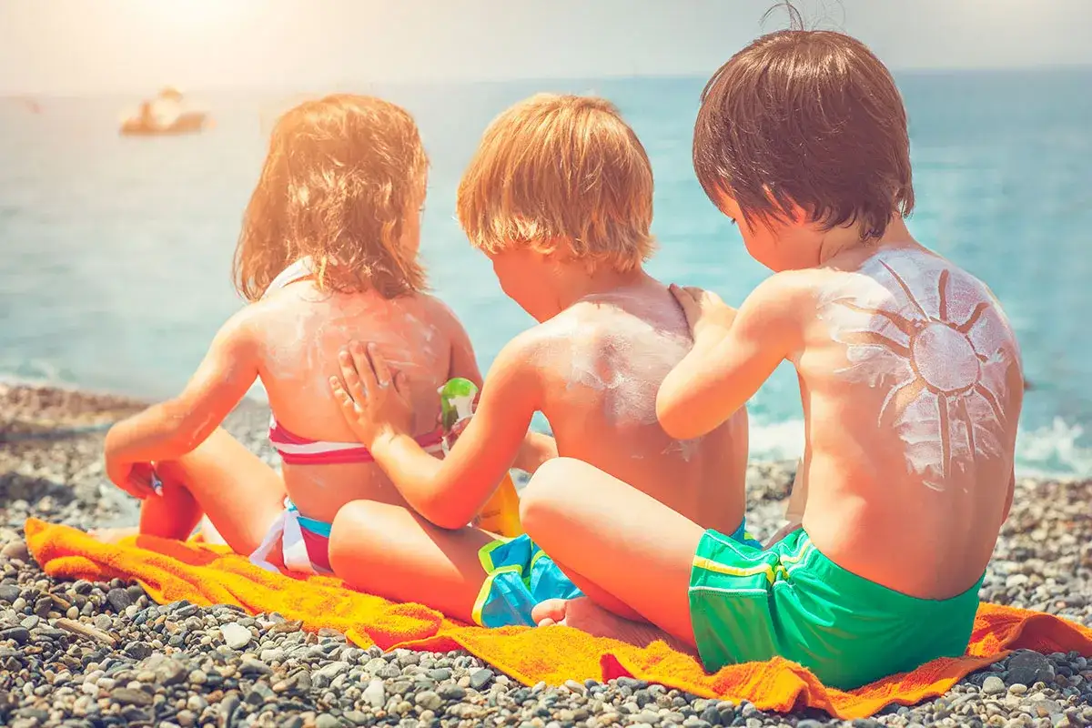 Bambini in spiaggia si spalmano la crema solare. Quanto dura la crema solare? Protezione solare disegnata a forma di sole sulla schiena di un bimbo.